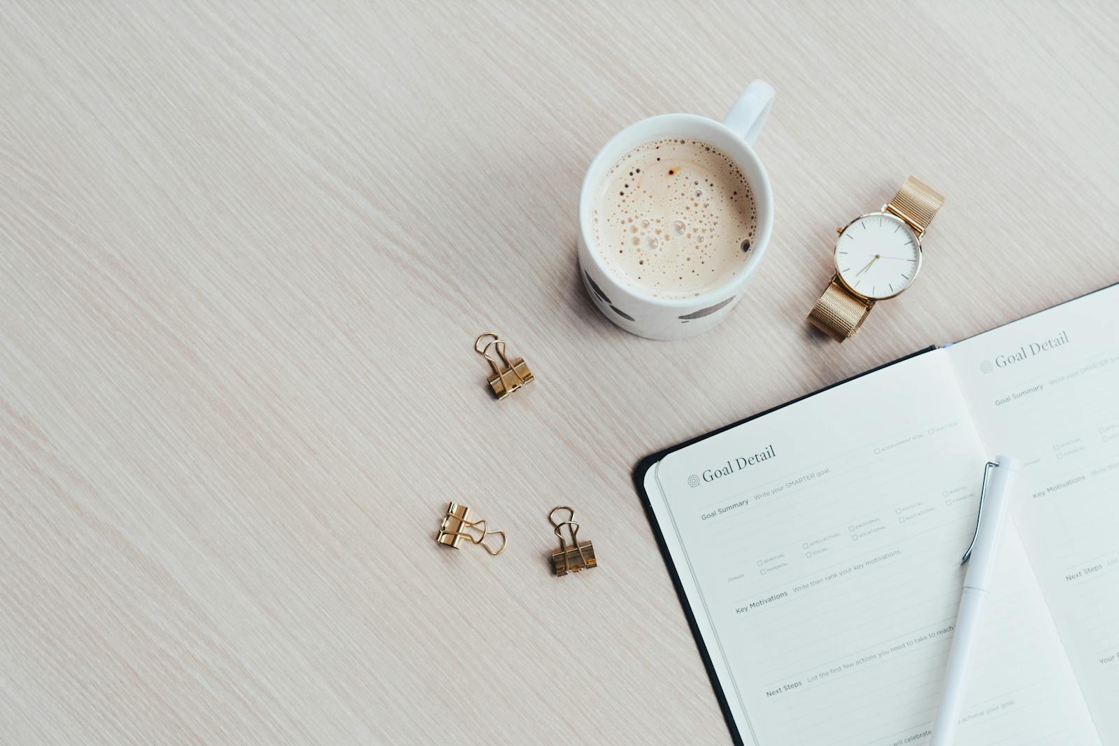 A minimalist desk arrangement featuring a coffee cup, notebook, pen, and wristwatch. for devotional Makings Plans